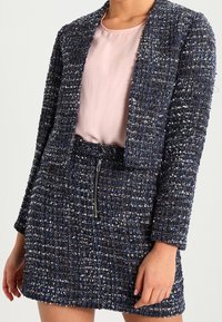 Texturovaný navy blazer a odpovídající sukně, s vystřihnutým designem, zipovými detaily a světle růžovou blůzou pod ním.