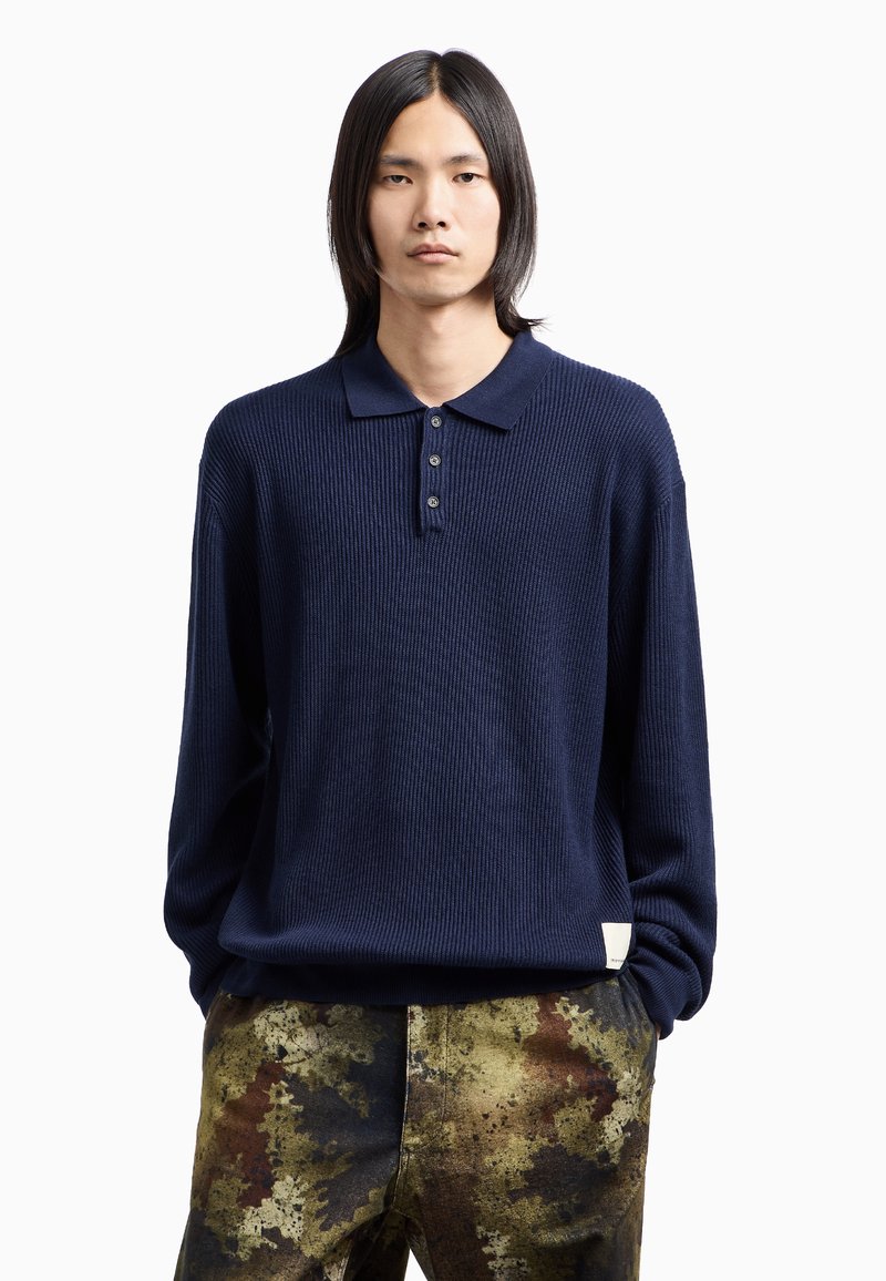 Polo shirt a coste blu navy con abbottonatura, abbinato a pantaloni tinti a trama multicolore con tonalità di marrone, oro e verde.