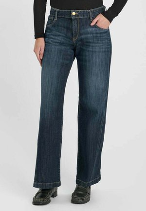 Personne portant un jean bleu foncé à jambes droites avec un bouton doré et des mocassins noirs, une main dans la poche, sur un fond clair uni.