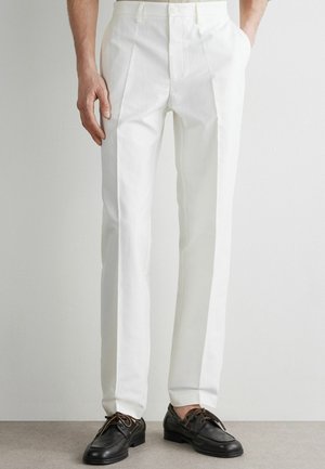 Mężczyzna w białych, dopasowanych spodniach i czarnych skórzanych mokasynach, stojący na tle jasnego, jednolitego tła.