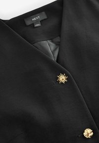Blazer nero con scollo a V, caratterizzato da un bottone dorato a raggiera e un tessuto strutturato. L'etichetta riporta la scritta "NEXT" con una fodera interna grigia.