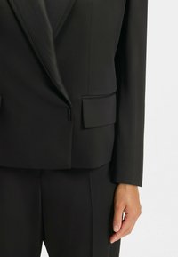 Zwarte op maat gemaakte blazer met een pieklapel, subtiele glans, gestructureerde schoudervullingen en zijzakken, gecombineerd met bijpassende broeken.
