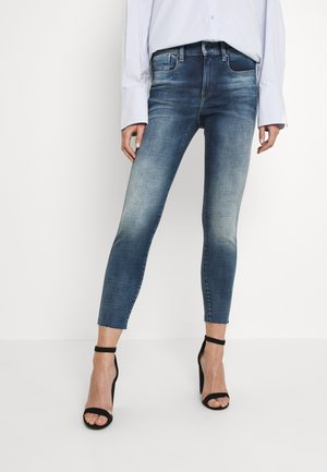 Kvinde iført falmede blå skinny jeans med frynser i kanten og sorte høje hælede sandaler, kombineret med en hvid langærmet skjorte.