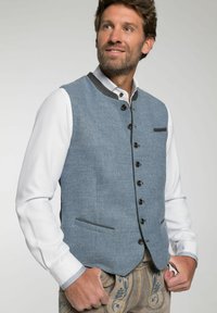 Blaues Leinenwesten mit hohem Kragen, schwarzen Knöpfen und einer Fronttasche; getragen über einem weißen Hemd mit gemusterten Manschetten.