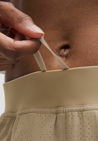 Beige athletic shorts with a smooth waistband and a drawstring; fabric features perforations for breathability. Hand adjusting the drawstring.