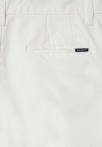 Pantaloni di cotone bianchi con una tasca posteriore con un'etichetta blu che recita "HACKETT." Texture liscia e linee pulite, con cuciture visibili.