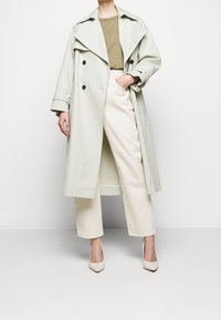 Jasnozielony trencz z czarnymi guzikami, szerokimi klapami i luźnym krojem, zestawiony z kremowymi spodniami o szerokim fasonie i beżowymi czółenkami na szpilce.