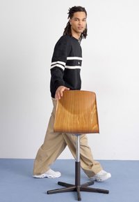Sgabello in legno con una seduta liscia e curvata in una tonalità calda, base in metallo elegante e quattro gambe nere. Modello che tiene lo sgabello, vestito con abbigliamento informale.