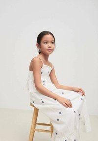 Robe blanche avec broderie bleue, bretelles spaghetti et ourlet festonné. Modèle assis sur un tabouret en bois sur un fond neutre.
