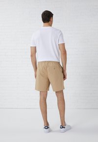 Beige Shorts mit elastischem Bund und einer Gesäßtasche, kombiniert mit einem schlichten weißen T-Shirt und weißen Sneakers, vor einer Backsteinmauer.