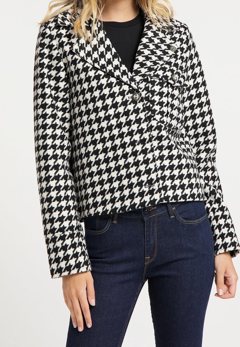 Femme portant une veste à motif pied-de-poule noir et blanc avec des boutons et un jean bleu foncé.