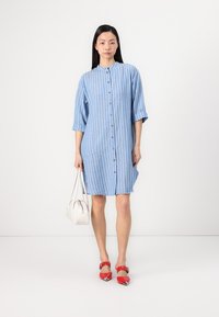 Vestido azul claro com riscas brancas verticais, bolsos laterais e mangas de meia, combinado com sapatos vermelhos e uma bolsa branca.