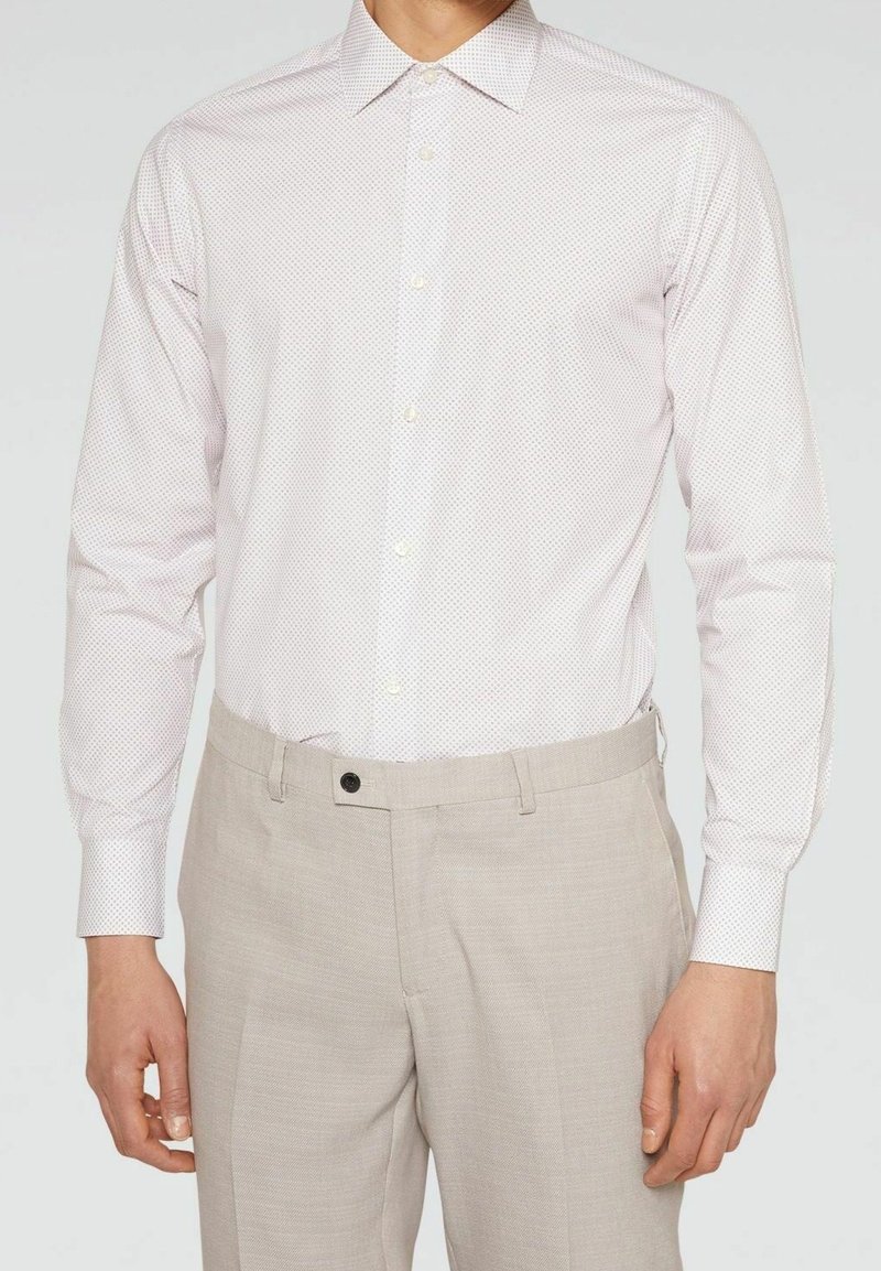 Homme portant une chemise blanche ajustée à manches longues avec un petit motif à pois, rentrée dans un pantalon beige clair de ville.