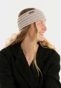 Grijze gebreide hoofdband met gestructureerde strepen en een klein leren logo-label, gedragen door een persoon in een zwarte gestructureerde jas tegen een lichte achtergrond.