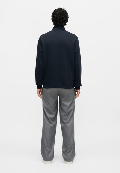 Άνδρας με κατσαρά μαλλιά που στέκεται με την πλάτη προς την κάμερα, φοράει μαύρο τοπ με μακρύ μανίκι, γκρι παντελόνι με φαρδιά μπατζάκια και λευκά παπούτσια.