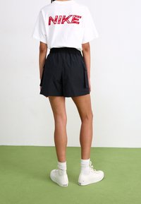 White t-shirt with red "NIKE" graphic on the back, paired with black shorts and white sneakers, set against a green floor.