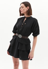 Schwarzes Kleid mit Puffärmel, mehrlagigem Rüschensaum und einem gebundenen Ausschnitt, tailliert mit einem braunen Gürtel mit einer runden silbernen Schnalle.