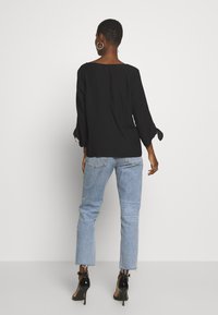 Top negro con mangas tres cuartos y detalles de lazo, combinado con jeans azul claro. La modelo lleva zapatos negros de tacón.