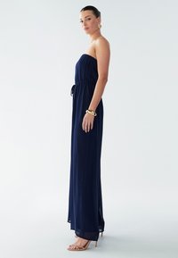 Temno modra brezrokavnikova maxi obleka z nabranci, elastiko v pasu in stranskimi razporki. Model nosi zlato nakit in nude sandale s trakovi.