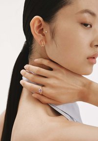 Pendientes de aro de oro y un anillo con piedra morada en una mano. Piel suave, tela gris claro y una coleta complementan la joyería.