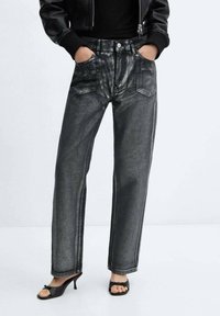 Jeans de mezclilla negros con acabado brillante, corte recto, bolsillos delanteros y detalles de costuras visibles. Combinados con sandalias negras de tacón.