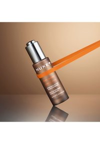 Brązowa szklana butelka z srebrną pompką, z pomarańczowym paskiem. Etykieta brzmi "NUXE Men Boost, serum wieloczynnościowe." Gładka tekstura.