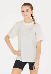T-shirt en coton beige clair à manches courtes et col rond, avec un petit logo vert sur la poitrine à gauche. Shorts noirs.