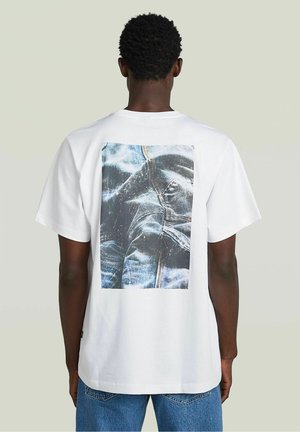 White cotton t-shirt featuring a large denim print on the back, with visible stitching details and a relaxed fit. Short sleeves included.