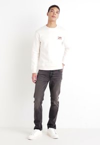 Sudadera de color claro con logo en el pecho, combinada con jeans oscuros y zapatillas blancas. Diseño simple y moderno con un ajuste relajado.