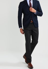 Man die een marineblauwe blazer aanpast, terwijl hij een gestreept overhemd, een bordeauxrode stropdas, zwarte broek, een bruine riem en bruine bedrijfsschoenen draagt, staand met één been gekruist.