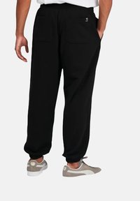 Pantalones de chándal negros con puños elásticos, que cuentan con dos bolsillos traseros. Combinados con zapatillas grises, mostrando un ajuste relajado y una textura de tela suave.