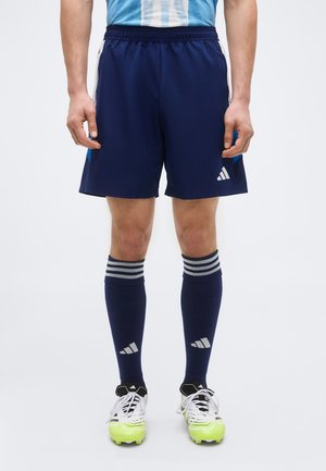 Athlète masculin portant un short de sport bleu, des chaussettes hautes assorties et des crampons blancs avec semelles jaune néon, debout devant un fond uni.