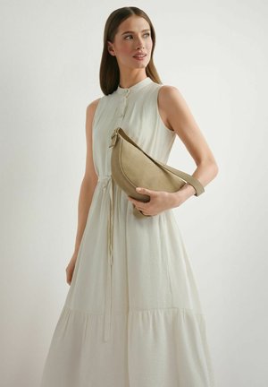 Kobieta w białej sukience bez rękawów trzymająca beżową torebkę w kształcie półksiężyca na jednolitym tle.