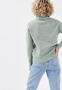 Suéter de lana verde claro con cuello alto y puños acanalados. Combinado con jeans azul claro, que muestran un ajuste relajado y una textura suave.