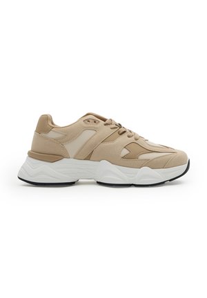 Zapatilla beige y marrón con parte superior de ante y malla mezclados, suela blanca gruesa con banda negra, y cierre de cordones, vista lateral sobre fondo blanco.