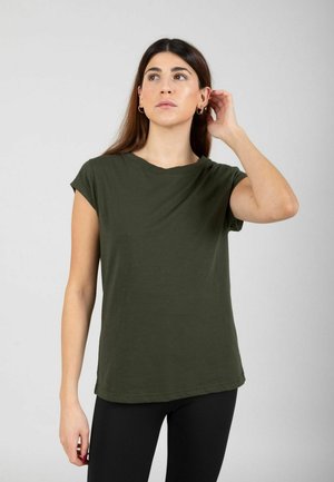 Femme aux longs cheveux foncés portant un t-shirt vert olive à manches courtes et un pantalon noir, touchant son oreille gauche, regardant légèrement vers le haut et à droite.
