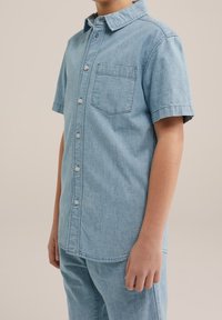 Světle modrá džínsová košile s krátkým rukávem, jednou kapsou na hrudi a viditelnými švy. Měkká tkanina a ležérní design.
