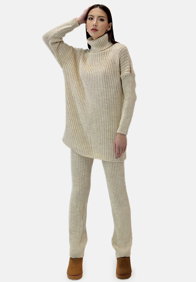 Elfenbenstrikoutfit med høj krave. Toppen er oversized med ribbet struktur, parret med tætsiddende ribbede bukser, komplementeret af brune ankelstøvler.