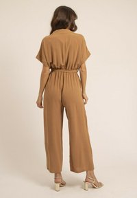 Jumpsuit marron, ample et décontracté, avec des manches courtes, une taille ceinturée et des jambes larges. Texture douce, design minimal et couleur unie.