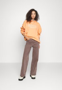 Felpa arancione con cappuccio e logo bianco, abbinata a pantaloni ampi marroni. Il modello indossa calzature chunky in bianco e nero.