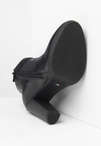 Botte noire en cuir à la cheville avec un talon texturé, des détails de fermeture éclair sur le côté et une surface lisse. La semelle présente un motif de bande de roulement pour une meilleure adhérence.
