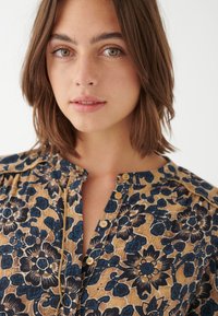 Blomstertryckt blus i beige, med blå och mörka blomsterdesigner. Den har en knappstängning framtill och en rund halsringning samt en strukturerad tyg.