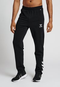 Zwarte joggingsbroek van zacht materiaal met een elastische tailleband, zijstrepen en een witte logo op de heup. Heeft een taps toelopende pasvorm.