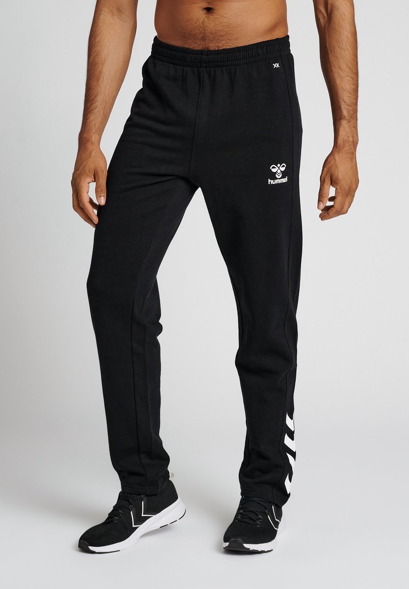 Zwarte joggingsbroek van zacht materiaal met een elastische tailleband, zijstrepen en een witte logo op de heup. Heeft een taps toelopende pasvorm.