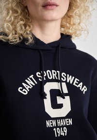 Personne blonde portant un sweat à capuche bleu marine avec l'inscription "GANT SPORTSWEAR", un grand "G" et "NEW HAVEN 1949" imprimés en blanc sur le devant.