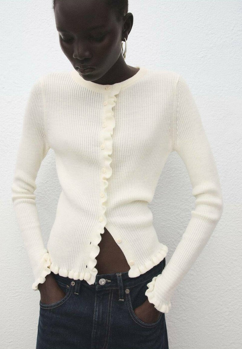 Cardigan crema a coste con bordi smerlati, chiusura con bottoni e maniche lunghe. Indossato con denim scuro. Design minimalista con texture morbida.