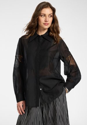 Nainen, jolla on päällään läpikuultava musta paita, jossa on pitsileikkauksia hihoissa, ja mustavalkoruudulliset leveät housut, katsoo sivulle käsi taskussa.