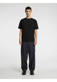 Melna T-krekla ar īsām piedurknēm un apaļu kakla izgriezumu, kopā ar tumši ziliem plašām biksēm. Modelis stāv priekšā neitrālam fona.