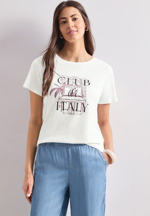 Femme aux longs cheveux bruns portant un T-shirt blanc avec le texte « Club di Italy » et un graphisme de bâtiment, associé à un pantalon bleu clair, debout contre un mur blanc.