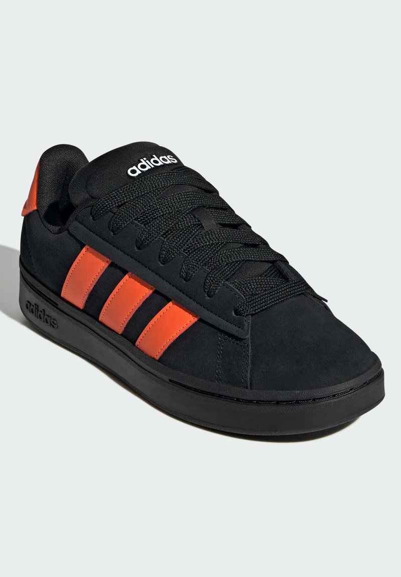 Sneakers Adidas noirs avec des rayures orange sur le côté, lacets noirs et semelle noire sur un fond gris clair.